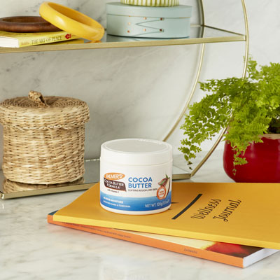 One of the best dry skin solutions, Palmer's Cocoa butter Formula Original Solid Jar on a table with wooden spoon scooping some out